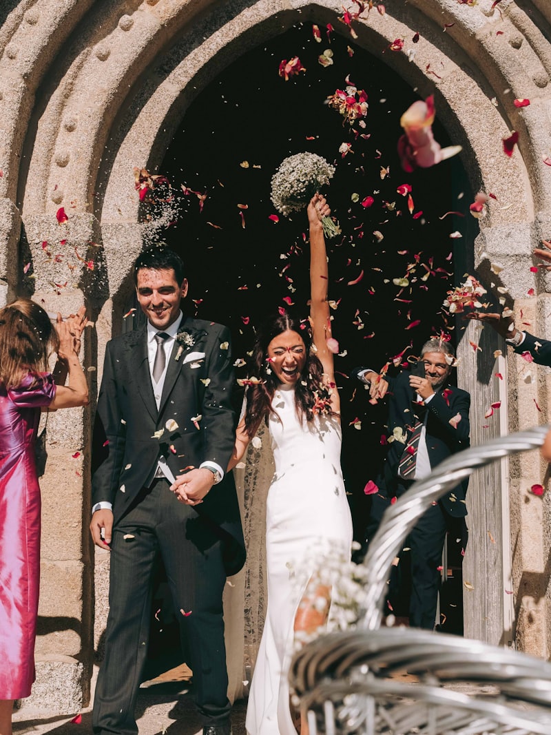 A couple leaving a stone church through a shower of petals, guests celebrating around them